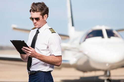 A male pilot stands outside of a corporate jet aircraft, using a modern digital tablet to update his flight plan and preflight checklist.